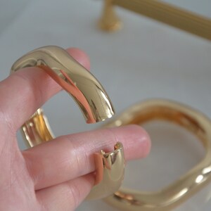 May include: Two gold-toned metal bangles, one with a wavy, organic shape and the other with a more rounded, oval shape.
