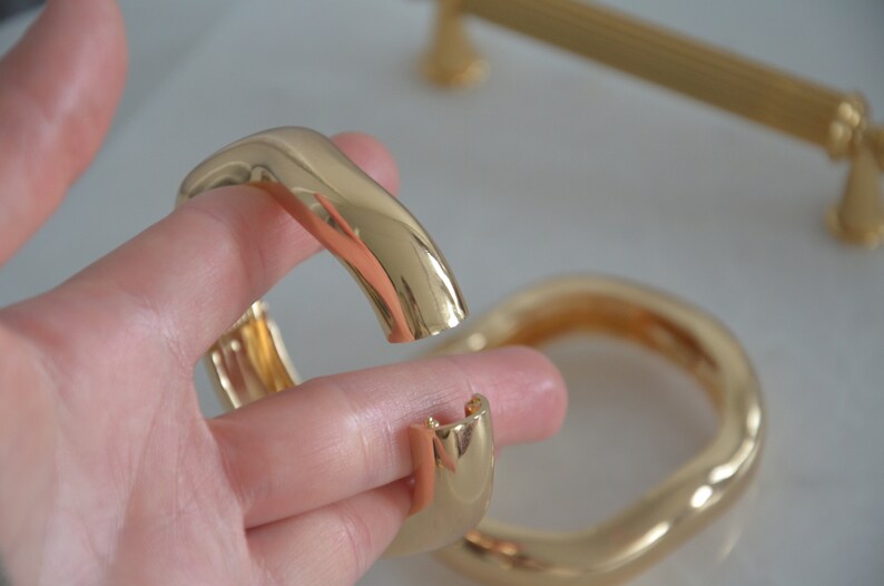 May include: Two gold-toned metal bangles, one with a wavy, organic shape and the other with a more rounded, oval shape.