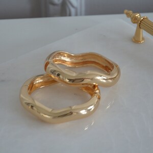 May include: Two gold-toned wavy rings, stacked on top of each other, on a white marble surface.