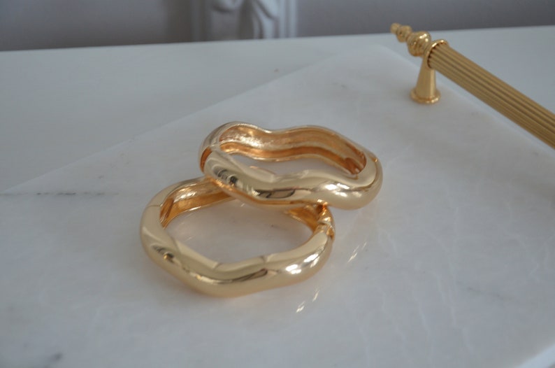 May include: Two gold-toned wavy rings, stacked on top of each other, on a white marble surface.