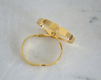 Könnte beinhalten: Zwei goldfarbene Armbänder mit einem Pflaster-Design. Die Armbänder sind offen und haben einen Scharnierverschluss.