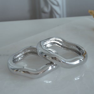 May include: Two silver, wavy, open-ended rings, stacked on top of each other. The rings are smooth and shiny.