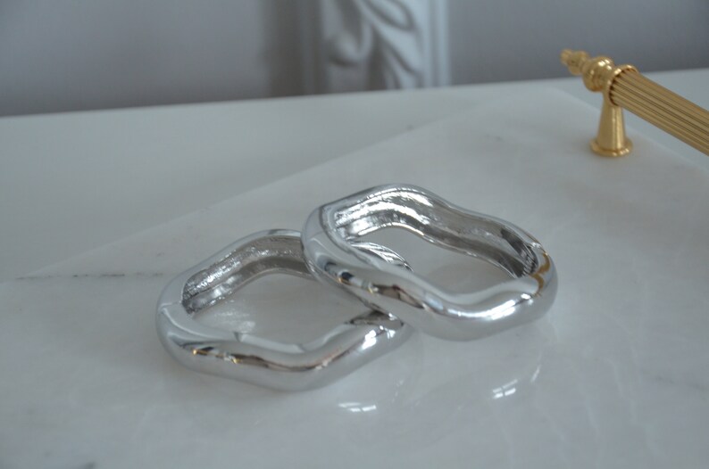 May include: Two silver, wavy, open-ended rings, stacked on top of each other. The rings are smooth and shiny.