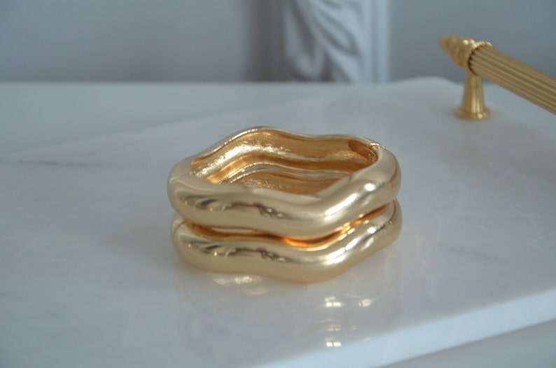 May include: Two gold-toned, wavy, open bangle bracelets stacked on top of each other.
