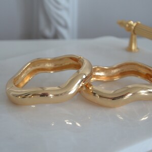 May include: Two gold-toned, wavy, open-ended bangle bracelets. The bracelets are made of a smooth, shiny metal and are stacked on top of each other.