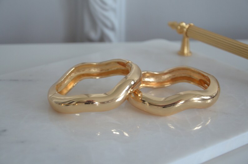 May include: Two gold-toned, wavy, open-ended bangle bracelets. The bracelets are made of a smooth, shiny metal and are stacked on top of each other.