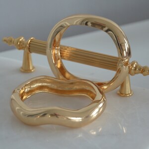 May include: Two gold-toned metal bracelets. One bracelet is a simple oval shape, the other is a wavy, organic shape.