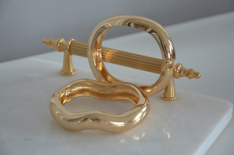 May include: Two gold-toned metal bracelets. One bracelet is a simple oval shape, the other is a wavy, organic shape.