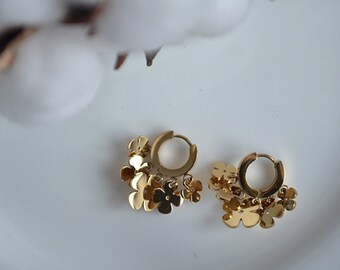May include: Gold-colored hoop earrings with dangling flower charms. The earrings are made of a shiny metal and feature small, detailed flower pendants. The earrings are displayed on a white surface, with a blurred cotton branch in the background.