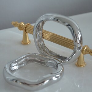 May include: Two silver cabinet door pulls, one with a gold bar and decorative ends. The pulls are on a white surface.