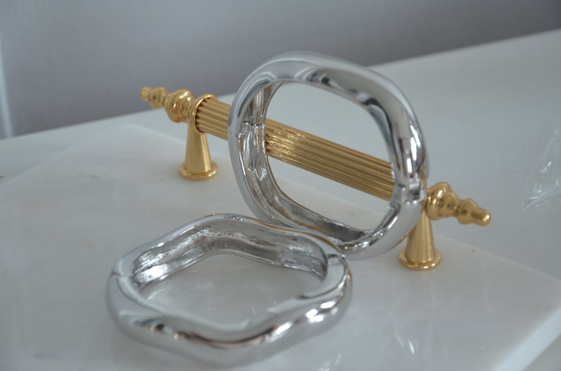 May include: Two silver cabinet door pulls, one with a gold bar and decorative ends. The pulls are on a white surface.