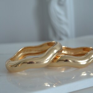 May include: Two gold-toned, wavy, open bangle bracelets. The bracelets are stacked on top of each other.