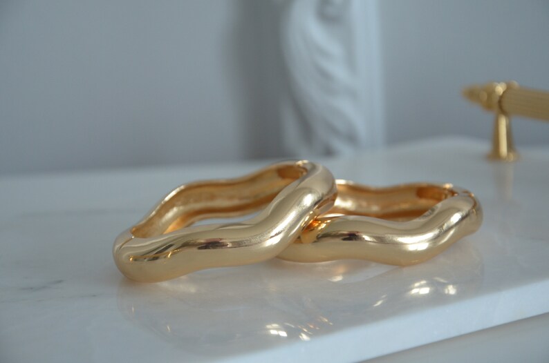 May include: Two gold-toned, wavy, open bangle bracelets. The bracelets are stacked on top of each other.