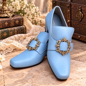 May include: A pair of light blue shoes with a square toe and a low block heel. Each shoe features a prominent decorative gold-toned metal buckle with an ornate, filigree-style scrollwork design centred on the vamp strap.