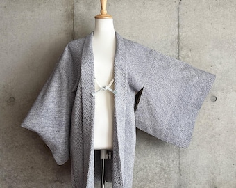 Könnte beinhalten: Eine texturierte Haori-Jacke in Grau und Weiß mit einem feinen, gesprenkelten Muster auf dem gesamten Stoff. Das Kleidungsstück hat weite, offene Ärmel und einen Frontverschluss, der mit einer hellgrauen Kordel befestigt ist, auf einer Puppe.