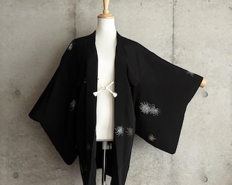 Könnte beinhalten: Eine schwarze Haori-Jacke mit einem Muster aus kleinen, verstreuten, mehrfarbigen Blumenmotiven in Blau-, Weiß- und Goldtönen. Das Kleidungsstück hat weite, lockere Ärmel und wird vorne mit einer weißen Kordel mit kleinen Quasten verschlossen.