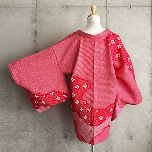 May include: A red kimono-style jacket with wide sleeves, displayed on a mannequin. The jacket features a textured weave and decorative patterns in red and white. The design includes floral and geometric motifs.