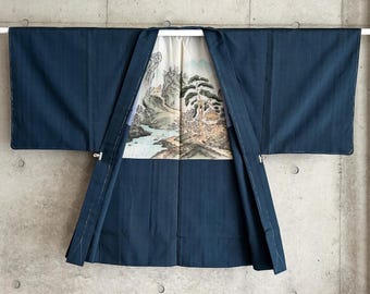 Könnte beinhalten: Eine dunkelblaue Haori-Jacke mit weiten Ärmeln und einem cremefarbenen Innenfutter, das eine detaillierte Landschaftsillustration mit Bergen, Kiefern und einem Fluss zeigt. Das Kleidungsstück hat einen strukturierten Kragen und sichtbare weiße Nähte.