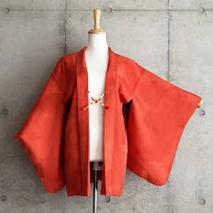 May include: A bright orange haori jacket featuring a subtle, tonal fan-shaped pattern throughout the fabric. The garment has wide, flowing sleeves and is secured at the front with a delicate, thin white and orange cord tie with small tassels.