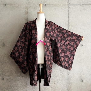 May include: A dark brown kimono-style jacket with a floral pattern in shades of pink and brown. The jacket is displayed on a mannequin and features wide sleeves and a tie closure. The garment is made of a textured fabric.