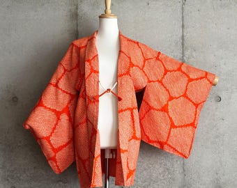 Könnte beinhalten: Eine orange-weiße Haori-Jacke mit einem sich wiederholenden geometrischen Muster aus großen, unregelmäßigen sechseckigen Formen. Das Kleidungsstück hat weite, lockere Ärmel, eine offene Vorderseite und einen kleinen weißen Kordelverschluss.