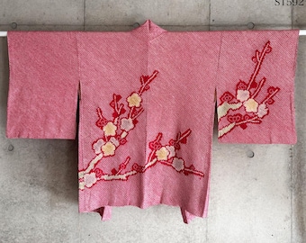 Könnte beinhalten: Ein rosa Kimono mit floralem Design. Der Kimono hängt an einer Betonmauer. Das florale Design zeigt rote Zweige mit weißen, gelben und rosa Blüten. Der Kimono hat weite Ärmel und einen Wickelstil.