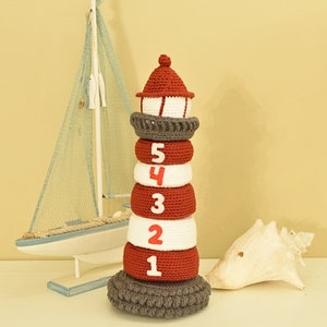 Puede incluir: Un juguete de faro de crochet rojo y blanco con una base gris. El faro tiene cinco anillos, cada uno con un número del 1 al 5.