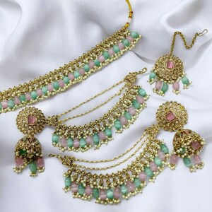 Puede incluir: Un conjunto de collar de oro con cuentas rosas y verdes, un par de pendientes a juego y un tikka. El collar tiene un dise&ntilde;o en capas con m&uacute;ltiples hileras de cuentas y cadenas.