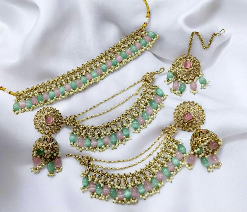 Puede incluir: Un conjunto de collar de oro con cuentas rosas y verdes, un par de pendientes a juego y un tikka. El collar tiene un dise&ntilde;o en capas con m&uacute;ltiples hileras de cuentas y cadenas.