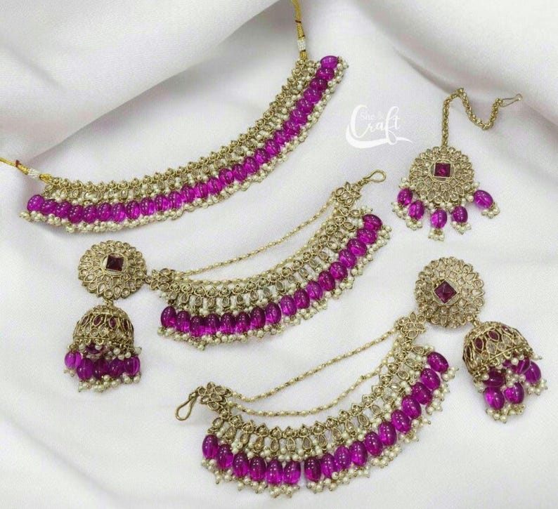 Puede incluir: Un conjunto de joyas doradas con cuentas rosas y piedras transparentes. El conjunto incluye un collar, un par de pendientes y un maang tikka.