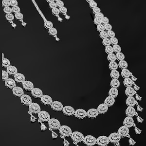 Puede incluir: Un collar plateado de dos hileras con piedras colgantes en forma de lágrima y pendientes a juego. El collar presenta una doble fila de piedras ovaladas.