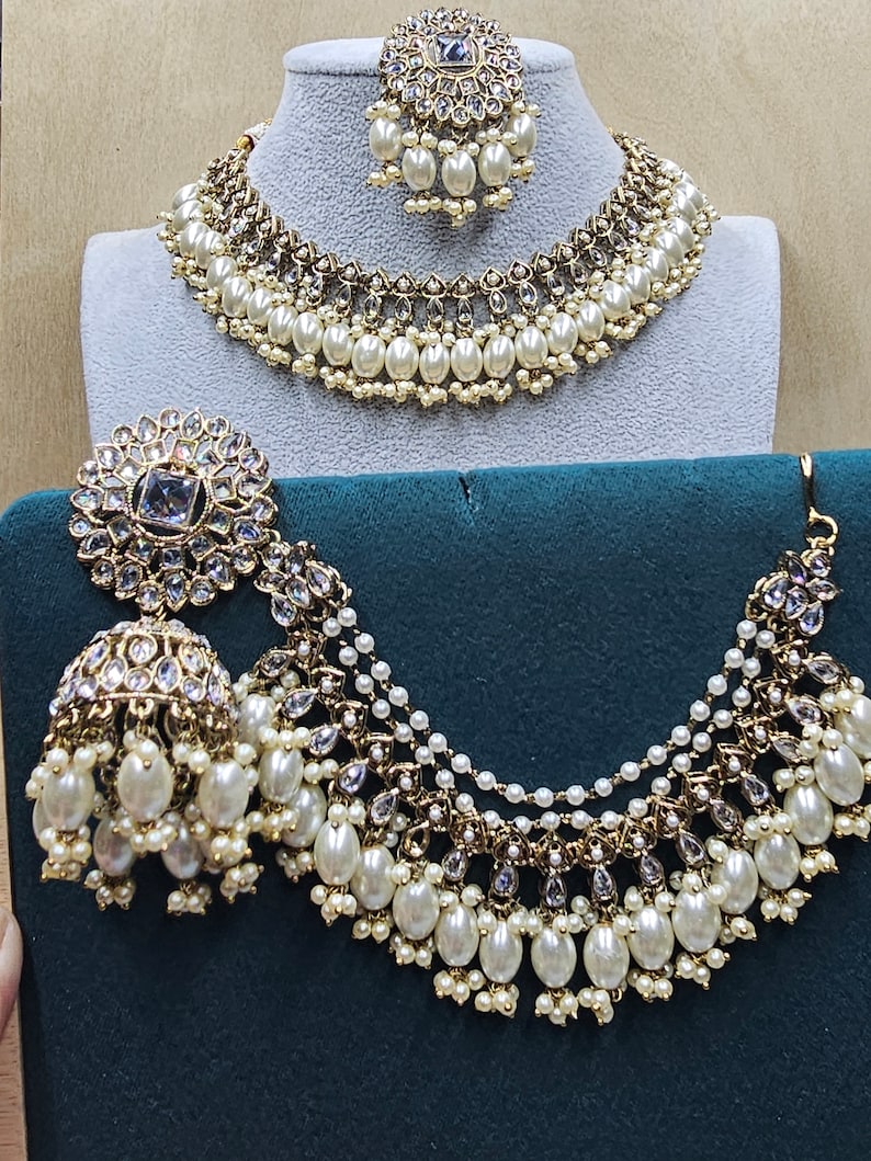 Puede incluir: Un conjunto de collar y pendientes de oro y perlas blancas. El collar tiene varias capas de perlas y cristales. Los pendientes son grandes y tienen forma de l&aacute;grima.