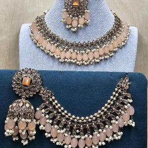Puede incluir: Un conjunto de collar y pendientes de color melocot&oacute;n con detalles dorados. El collar tiene tres filas de cuentas y los pendientes son de forma de l&aacute;grima.