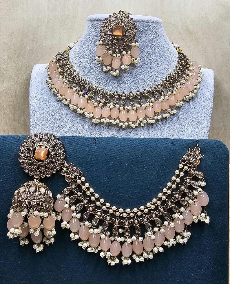Puede incluir: Un conjunto de collar y pendientes de color melocot&oacute;n con detalles dorados. El collar tiene tres filas de cuentas y los pendientes son de forma de l&aacute;grima.