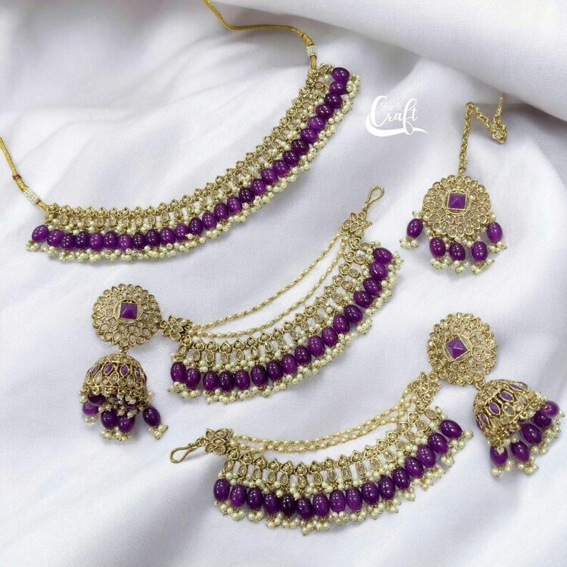 Puede incluir: Un conjunto de joyas de cuentas moradas y doradas con un collar, pendientes y un maang tikka. Las joyas presentan cuentas moradas redondas y detalles dorados.