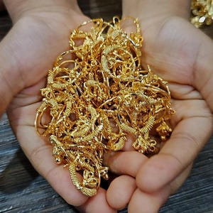 Op de afbeelding: Een handvol gouden sieraden, waaronder kettingen en armbanden, wordt in open handen gehouden. De sieraden hebben ingewikkelde ontwerpen en een heldere, glanzende afwerking. De achtergrond is een donker houten oppervlak.