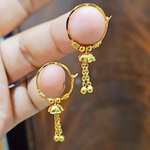 May include: A pair of gold-colored earrings. Each earring features a circular design with a pink sphere at the center. Below the sphere hangs a small, bell-shaped element with dangling chains and small, round accents.