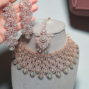 May include: A rose gold-toned jewelry set featuring a necklace, earrings, and a headpiece. The necklace and earrings have a floral design with clear and light green stones. The earrings are held by a hand.