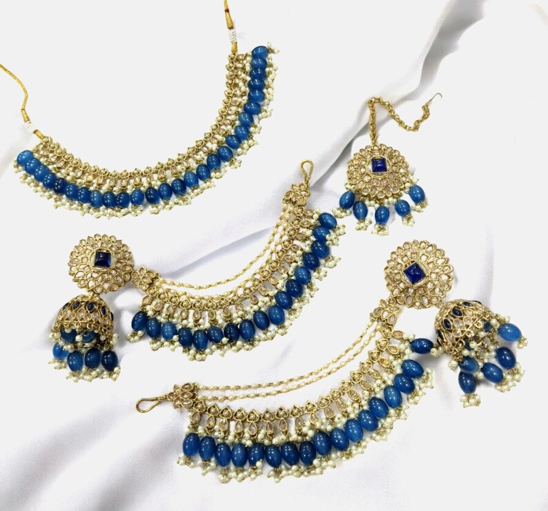 Puede incluir: Un conjunto de joyas de novia indias azul y dorado con un collar, pendientes y un maang tikka. El conjunto presenta cuentas azules, detalles dorados y piedras brillantes.