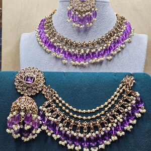 Puede incluir: Un conjunto de collar y pendientes de novia indio en oro y morado. El collar presenta un dise&ntilde;o en capas con cuentas moradas y detalles dorados. Los pendientes tambi&eacute;n est&aacute;n adornados con cuentas moradas y detalles dorados.