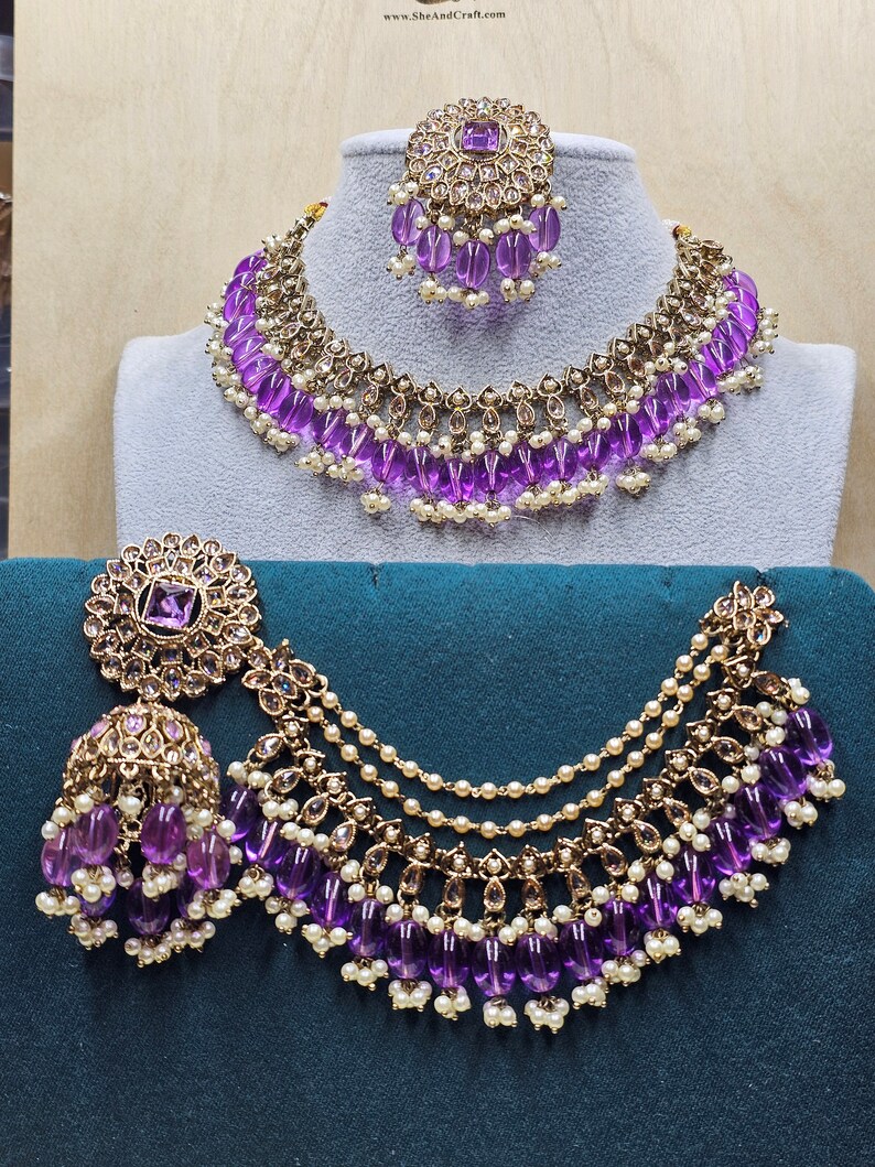 Puede incluir: Un conjunto de collar y pendientes de novia indio en oro y morado. El collar presenta un dise&ntilde;o en capas con cuentas moradas y detalles dorados. Los pendientes tambi&eacute;n est&aacute;n adornados con cuentas moradas y detalles dorados.