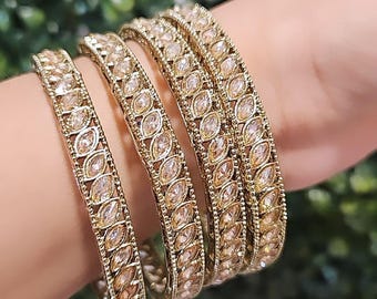 May include: Four gold bangles with clear rhinestone embellishments. The bangles are stacked on a person's wrist.