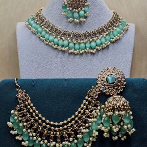 Puede incluir: Un conjunto de collar y pendientes de novia indio en verde y dorado. El collar tiene varias hileras de perlas y cuentas verdes, mientras que los pendientes son grandes y ornamentados.