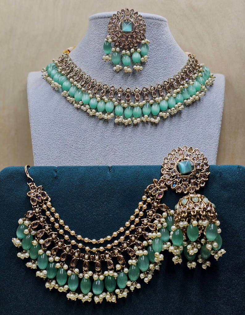 Puede incluir: Un conjunto de collar y pendientes de novia indio en verde y dorado. El collar tiene varias hileras de perlas y cuentas verdes, mientras que los pendientes son grandes y ornamentados.