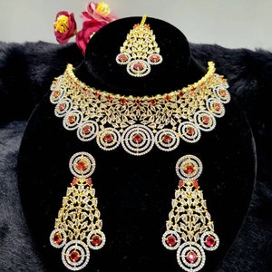 May include: A gold-tone bridal jewellery set with a choker necklace, earrings, and a maang tikka. The set features clear cubic zirconia stones and red accents.