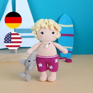 Puede incluir: Muñeca de ganchillo con pelo rubio rizado, que lleva pantalones cortos rosas con diseños de flores blancas y un collar de conchas. La muñeca sostiene un tiburón gris de ganchillo. Una tabla de surf azul está en el fondo.