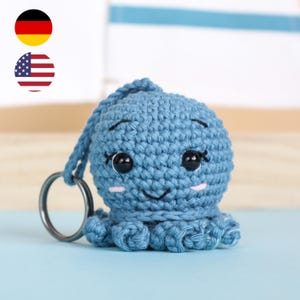 Può includere: Un portachiavi a forma di polpo blu all'uncinetto fatto a mano. Il polpo ha un viso sorridente, occhi neri e guance rosa. Presenta otto tentacoli arricciati e un portachiavi argentato.