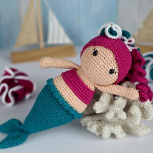 May include: A crocheted mermaid doll with pink hair, a pink top, and a teal tail. The doll is lying on a white coral reef.
