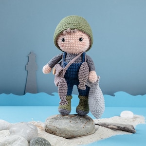 Może przedstawiać: Pluszowa lalka amigurumi w postaci rybaka, wykonana na szydełku. Lalka ma na sobie zielony kapelusz, niebieskie ogrodniczki i zielone buty. Lalka trzyma wędkę i worek z rybami.