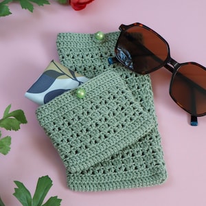 May include: Sage green crochet sunglasses case with a button closure, holding a patterned fabric. Tortoiseshell sunglasses with brown lenses rest beside the case. The items are on a pink surface, with red flowers and green leaves.